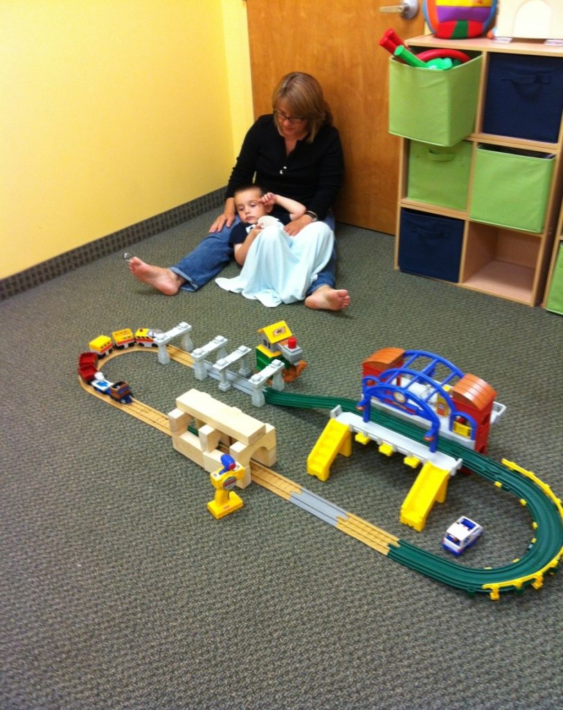 Julie sits on the floor with a child holding a blanket. Julie and the child are in the corner of the room, with a shelf to their left. An assembled train is on the floor before them.