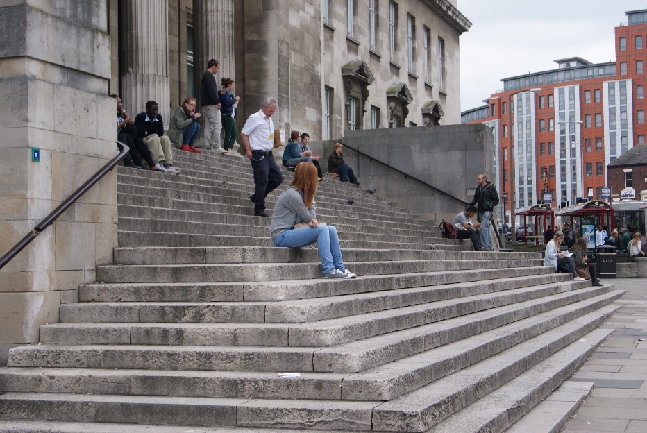 The front of a limestone building is shown with a wide, broad staircase of the same limestone in front. Multiple people are either walking or sitting on the stairs.