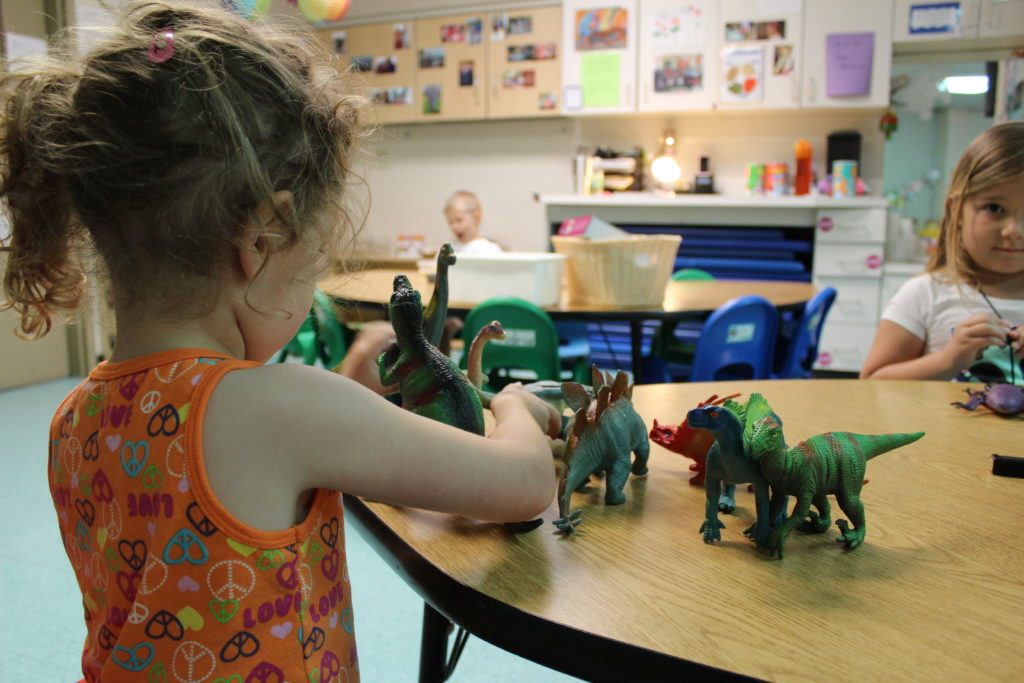 A girl stands with her back to the camera playing with several plastic dinosaurs at a table. Several other children in the room are visible in the background.