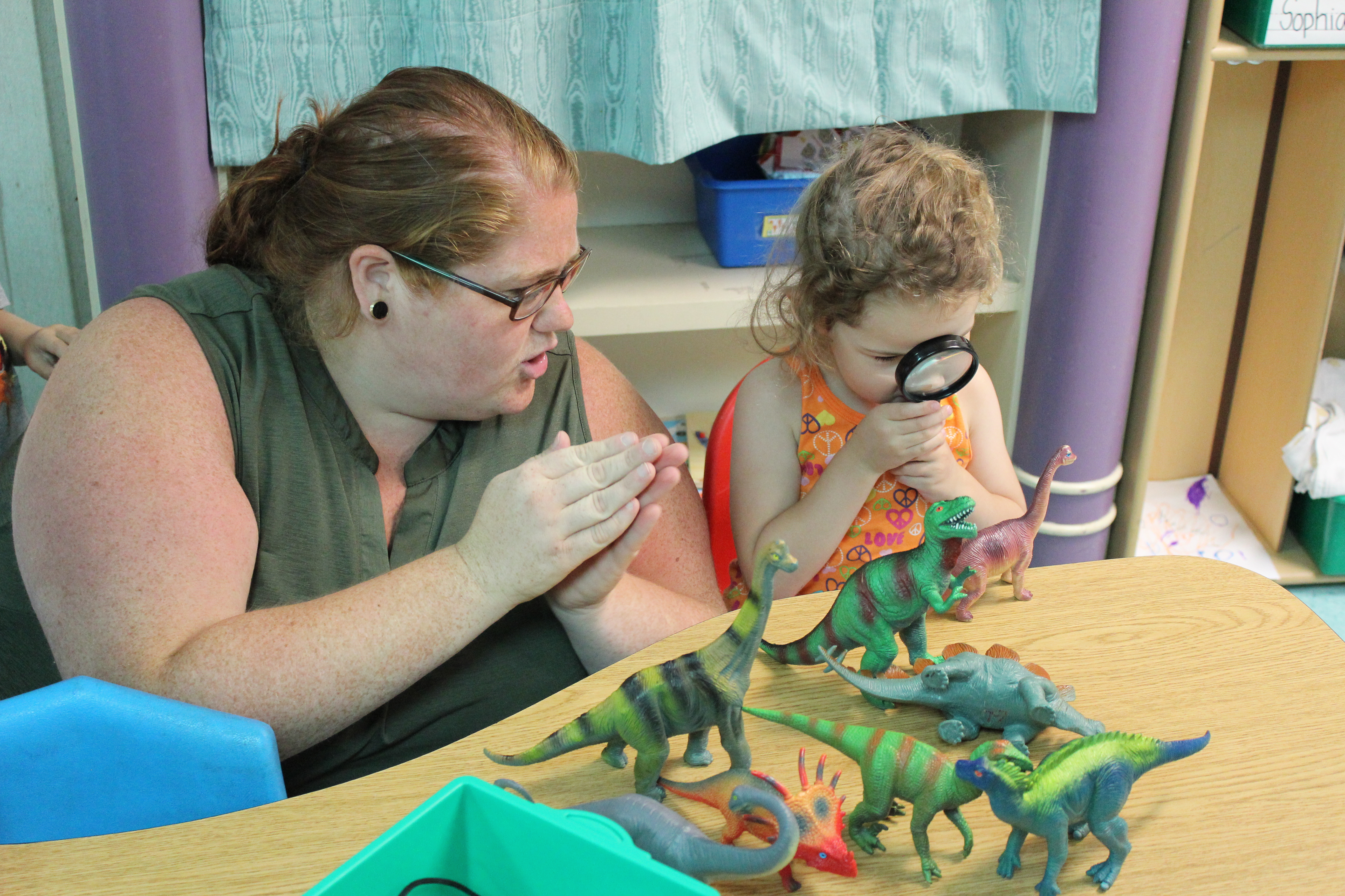 Teacher sitting next to a child at a table. The child has a magnifying glass in her hand and is looking at plastic dinosaurs.