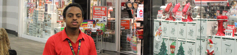 A man wearing a red shirt and a lanyard stands in front of a kitchen store in the mall.