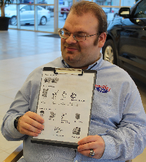 A man sits in a car dealership show room, holding a clipboard.