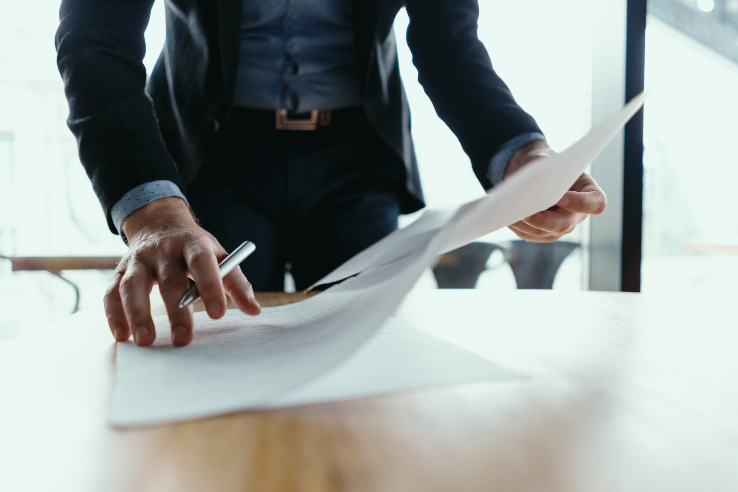 Close up hands signing documents in a modern office with window in background. Pen in hand, papers on the wooden desk, futuristic background.