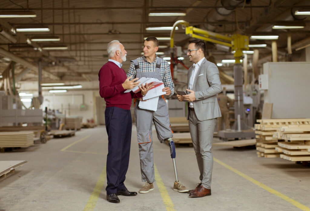 Three men discussing papers in a warehouse. The man in the middle has a prosthetic leg.