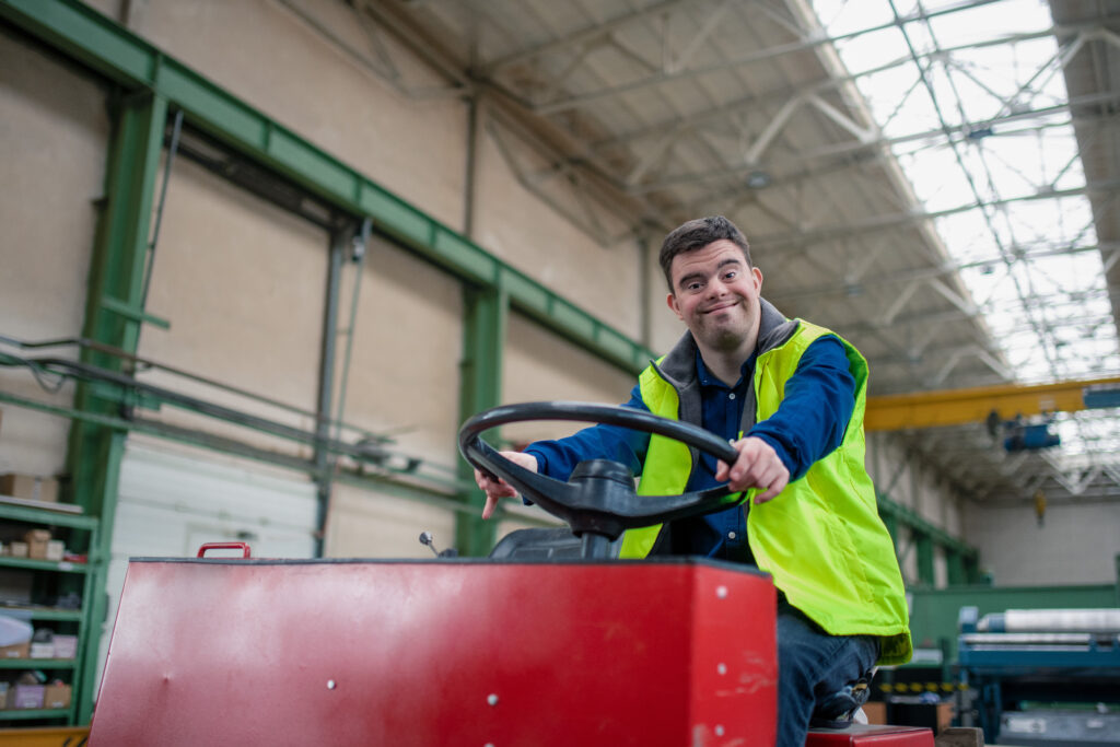 A man sitting at the stearing wheel of a piece of equipment in a warehouse wearing a yellow vest
