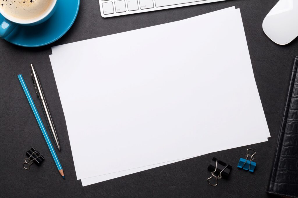 Blank piece of white paper on a desktop surrounded by a coffee cup, keyboard, binder clips, and writing instruments.