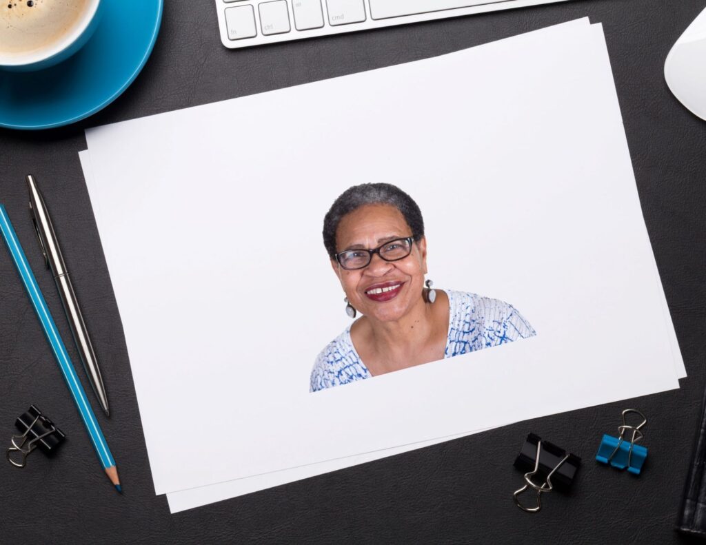 Piece of white paper with an image of an older woman wearing glasses on it,  on a desktop surrounded by a coffee cup, keyboard, binder clips, and writing instruments.