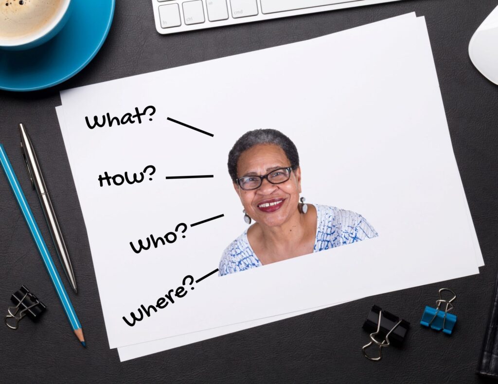 Piece of white paper with an image of an older woman wearing glasses on it.  Words “what”, “how”, “who”, and “where” are written to the left of the woman with lines pointing from the words to the woman.  Paper is on a desktop surrounded by a coffee cup, keyboard, binder clips, and writing instruments.