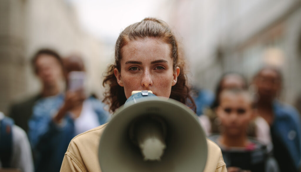 Female activist protesting with megaphone during a strike with group of demonstrator in background. Woman protesting in the city.