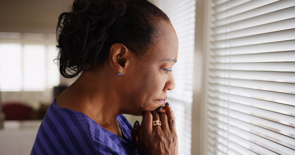 Woman looking sadly out the window holding her hands below her chin.