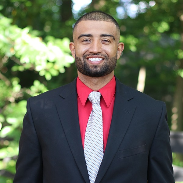 An early career mixed man with dark hair and brown skin wears a red shirt, white tie and dark suit. He is standing outside and smiling into the camera.