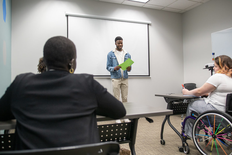 A student stands before a class presenting a paper. Seated, you can see two other students, one is a power wheelchair user.